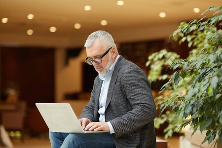 Portrait of contemporary senior businessman using laptop while working in office hall, copy spaceの写真素材