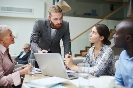 Portrait of successful young businessman leading meeting with employees in office and pointing at laptop screen, copy spaceの写真素材