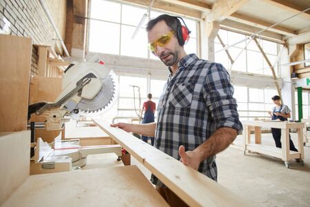 Waist up portrait of mature carpenter sawing wood in industrial workshop, copy spaceの写真素材