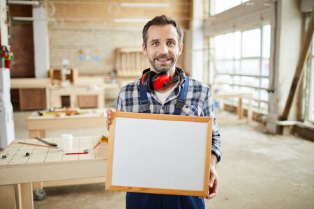 Waist up portrait of smiling mature carpenter holding blank frame and looking at camera, copy spaceの写真素材