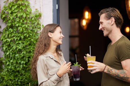 Side view portrait of smiling young woman talking to man outdoors standing by cafe door, both holding cold refreshing drinks, copy spaceの写真素材