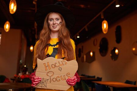 Waist up portrait of red haired woman wearing Halloween costume posing with Hocus Pocus sigh during party in nightclub, copy spaceの写真素材