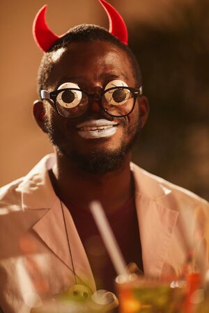 Portrait of African man wearing Halloween costume posing as devil with googly eyes during party,...