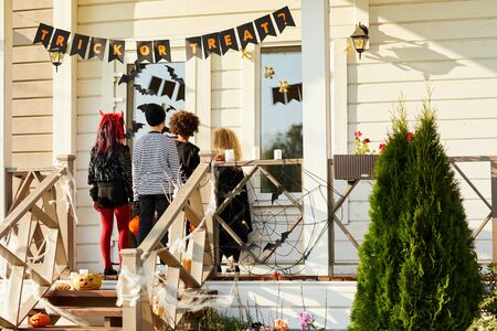 Back view of children trick or treating on Halloween, kids standing on porch knocking on doors of decorated house, copy spaceの写真素材