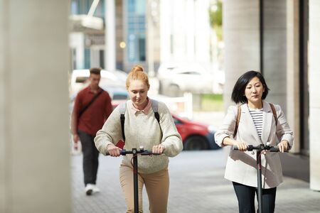 Portrait of two women riding electric scooters while commuting in urban city, copy spaceの写真素材