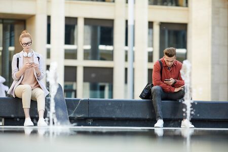 Full length portrait of two contemporary young people using smartphone sitting on bench in urban setting, copy spaceの写真素材