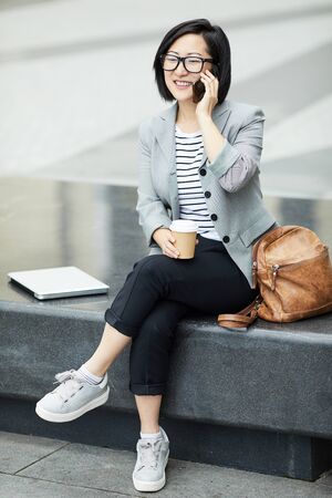 Full length portrait of modern Asian woman speaking by smartphone outdoors and smiling happily, scene in urban settingの写真素材