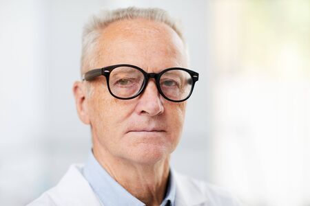 Head and shoulders portrait of white haired senior doctor wearing glasses and looking at camera while posing in office, copy spaceの写真素材
