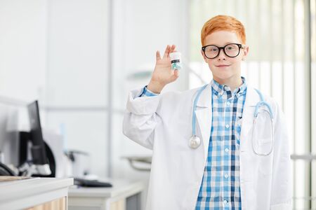 Waist up portrait of cheerful little doctor wearing white coat holding pill bottle and smiling happily at camera, copy spaceの写真素材