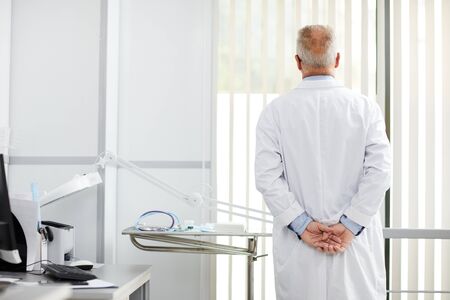 Back view portrait of pensive senior doctor looking at window while standing in office of modern clinic, copy spaceの写真素材