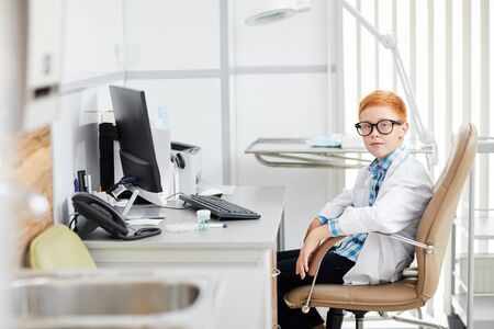 Portrait of cute red haired boy posing as doctor wearing white coat while sitting at desk in office and looking at camera, copy spaceの写真素材
