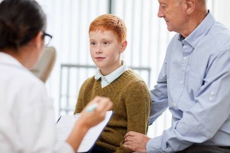 Portrait of cute teenage boy visiting doctor with father and looking at nurse filling patients form in waiting room, copy spaceの写真素材