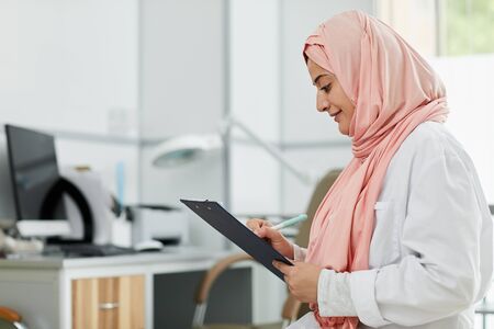 Side view portrait of Middle-Eastern woman wearing hijab working as nurse in medical clinic and writing on clipboard, copy spaceの写真素材