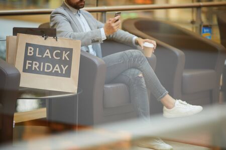 Cropped portrait of fashionable young man using smartphone sitting in comfortable armchair in mall, shopping bags with Black Friday inscription beside him, copy spaceの写真素材