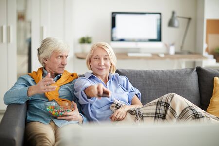 Portrait of modern senior couple watching TV together, focus on frustrated husband looking at wife...