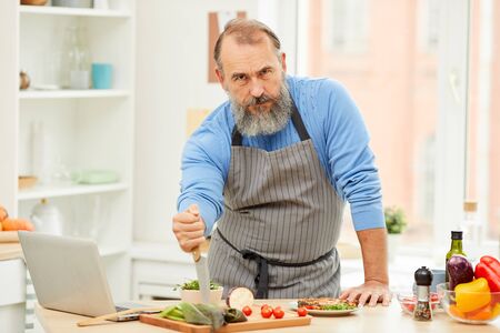 Portrait of tough bearded man holding knife and looking at camera while cooking meat in kitchen, copy spaceの写真素材