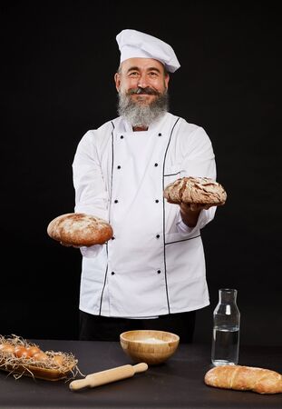 Portrait of bearded baker holding fresh bread smiling happily at camera while posing against black backgroundの写真素材