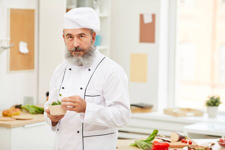 Waist up portrait of bearded senior chef looking at camera holding bowl with fresh herbs while standing in restaurant kitchen, copy spaceの写真素材