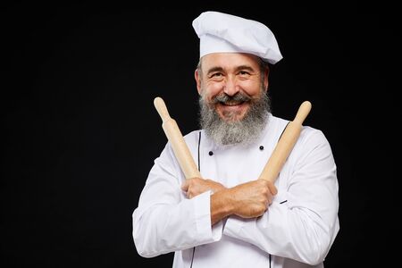 Waist up portrait of cheerful bearded baker holding two rolling pins while standing against black background and smiling at camera, copy spaceの写真素材