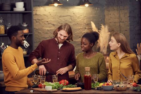 Multi-ethnic group of young people enjoying dinner together standing at table in modern interior and chatting, copy spaceの写真素材