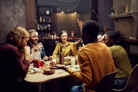 Group of young friends playing guessing game while sitting at table during dinner party in dark room, copy spaceの写真素材