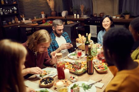 Multi-ethnic group of friends enjoying delicious dinner together sitting at table in dimly lit room, focus on smiling Asian womanの写真素材