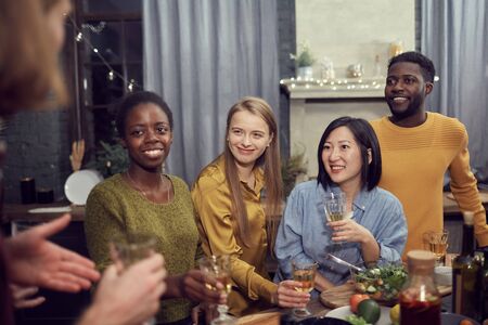 Multi-ethnic group of young people enjoying party standing at table in modern interior and listening to friend telling storyの写真素材
