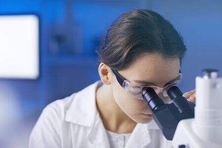 Closeup portrait of young female scientist looking in microscope while doing research in medical...