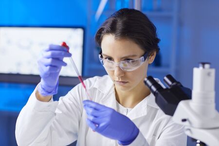 Portrait of young female scientist preparing blood test sample using dropper while working in...