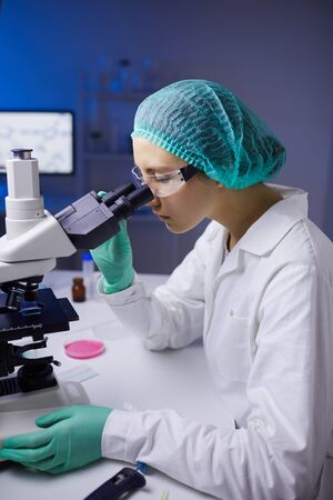 Side view portrait of young woman looking in microscope while working on medical research in...