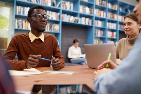 Portrait of smart African-American man smiling happily while studying with group of students in...