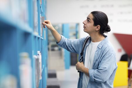 Side view portrait of Latin-American student choosing books standing by shelves in college library,...