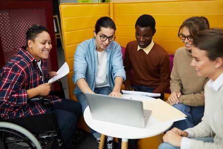 Multi-ethnic group of students using laptop while discussing team project during meeting in college...
