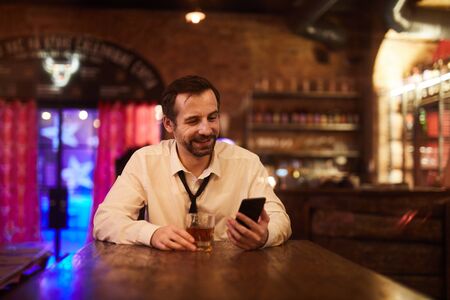 Portrait of mature businessman drinking alcohol in bar after work and smiling looking at smartphone screen, copy spaceの写真素材