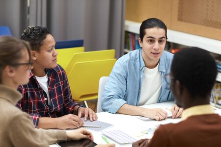 Multi-ethnic group of students sitting at table in college library and discussing project while...