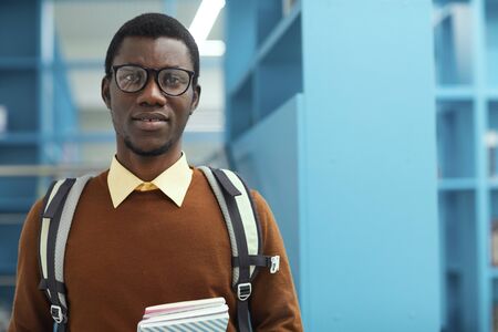 Waist up portrait of smart African-American student holding book standing in college library, copy spaceの写真素材