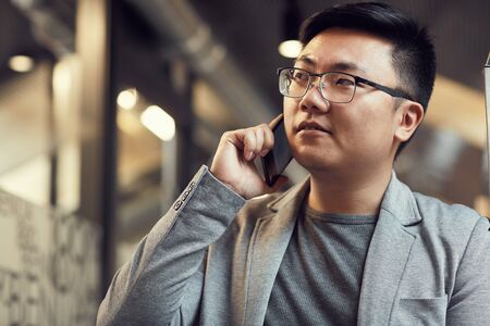 Portrait of successful Asian businessman speaking by phone while standing in modern office interior, copy spaceの写真素材