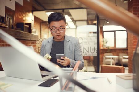 Portrait of young Asian entrepreneur using smartphone while working at desk in modern office, copy spaceの写真素材