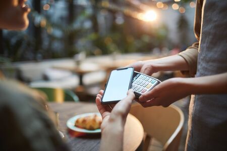 Close up of young woman paying via NFC in cafe, focus on female hands holding smartphone with blank screen to banking terminal, copy spaceの写真素材