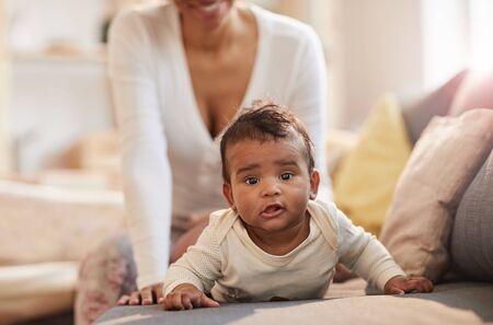 Portrait of cute African-American baby looking at camera while lying on couch with young mother...