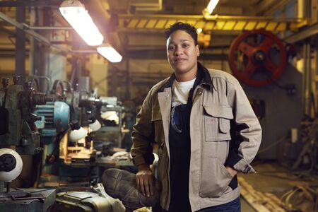 Waist up portrait of contemporary mixed-race woman posing in factory workshop at looking at camera,...