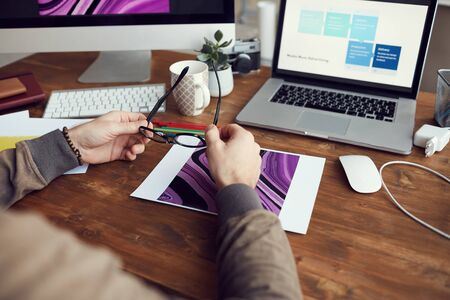 Close up of unrecognizable male designer working with images while sitting at desk in home office and holding glasses, copy spaceの写真素材