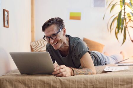 Portrait of contemporary adult man lying on bed and using digital tablet while enjoying entertainment videos at home, copy spaceの写真素材