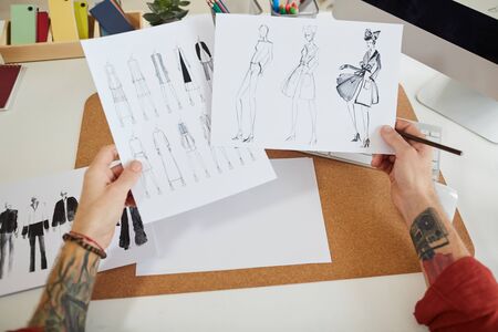 Close up of tattooed male hands holding fashion sketches over workplace in design studio, copy space