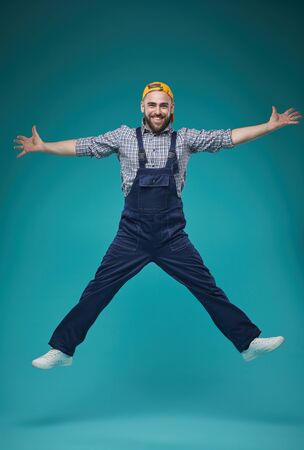 Vertical studio full shot of cheerful young adult guy in workwear having fun jumping, blue turquoise backgroundの写真素材
