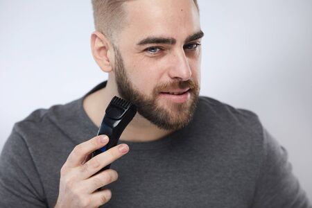 Head and shoulders studio portrait shot of stylish young man grooming his beard using trimmer...