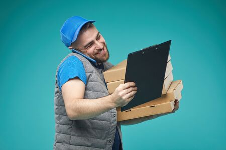 Horizontal medium studio shot of delivery man holding pizza boxes, talking on phone and checking order listの写真素材