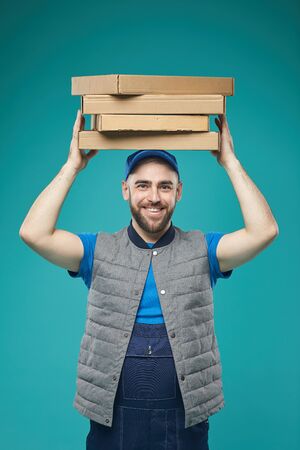 Vertical studio portrait of cheerful food delivery worker holding pizza boxes above his head looking...