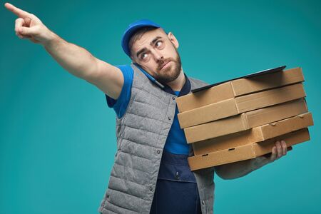 Horizontal medium studio shot of food delivery male worker holding pizza boxes, talking on phone and pointing at somethingの写真素材