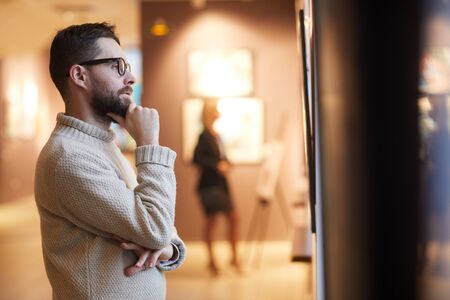 Side view portrait of mature bearded man looking at paintings while enjoying exhibition in modern gallery or museum, copy spaceの写真素材